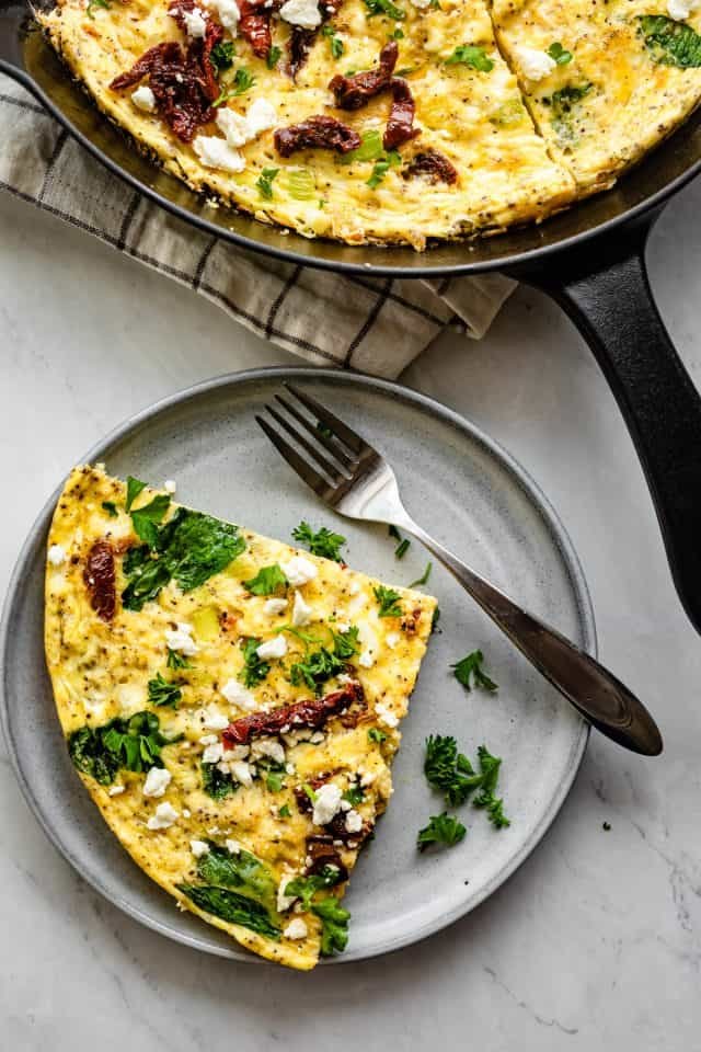 A slice of spinach and vegetables frittata topped with feta cheese and cilantro, served on a small blue plate with a fork on a table with skillet of frittata in the background. 