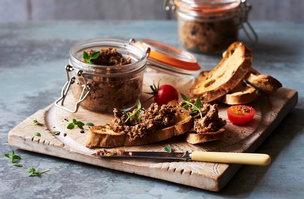 Homemade vegan mushroom Pâté in a small jar spread on brushetta on a chopping board with 2 small fresh tomatoes