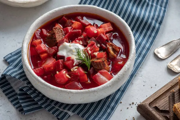 homemade borscht soup with beef served in a white bowl with a blue and whiite striped table cloth on a table garnish with sour cream and dill